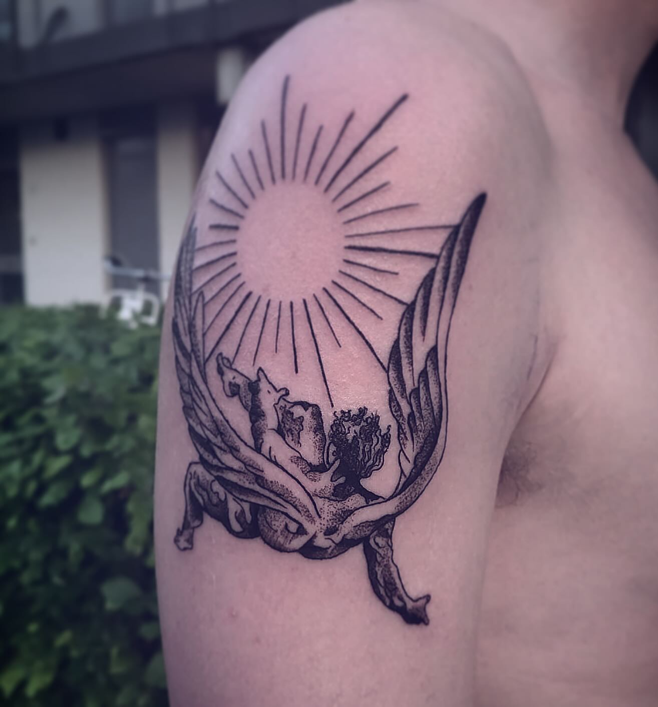 Fine line tattoo van een zon met stralen en een gevleugelde figuur op een bovenarm